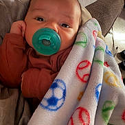 Richie joined the competition — help win amazing prizes! baby, pacifier, blanket, peace_sign, soccer_ball, pillow, couch, infant, clothing, face, hand, comfort, indoor, soft_texture, resting, cute, portrait, child, snuggle, warm