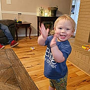 Elijah is registered to the contest to win money with this photo: toddler, child, clapping, smiling, blue_shirt, big_brother_shirt, pajama_pants, barefoot, wooden_floor, living_room, table, brick_fireplace, plant, toy, furniture, happy, indoor, person, young_child, home