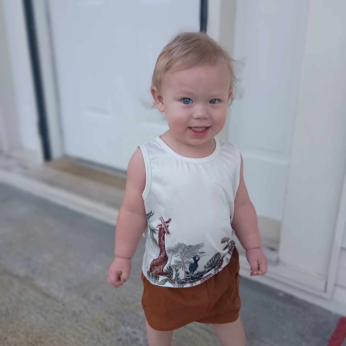 Elijah is registered to the contest to win money with this photo: animal_print, blue_eyes, casual_clothing, child, concrete_floor, cute, door, giraffe, happy, outdoor, person, playful, portrait, sandals, shorts, sleeveless_shirt, smiling, standing, toddler, young_child