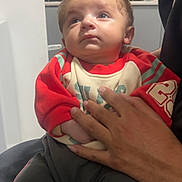 Hannan joined the competition — help win amazing prizes! adult_hand, baby, black_pants, casual_clothing, child, curious_expression, hand, holding, indoor, infant, kitchen, light_switch, looking_up, person, portrait, red_clothing, sitting, sweater, tile_wall, young_child