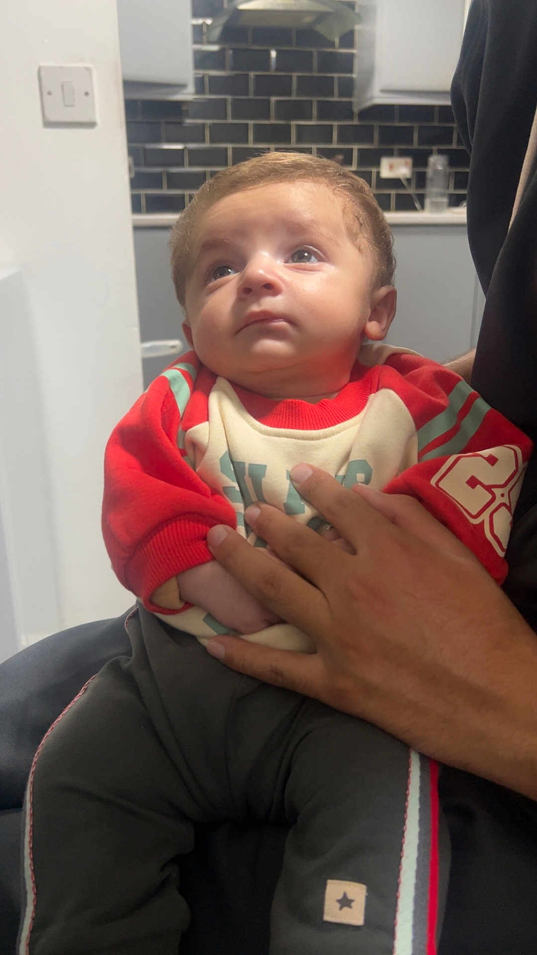 Hannan joined the competition — help win amazing prizes! adult_hand, baby, black_pants, casual_clothing, child, curious_expression, hand, holding, indoor, infant, kitchen, light_switch, looking_up, person, portrait, red_clothing, sitting, sweater, tile_wall, young_child