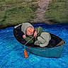 newborn, baby, sleeping, boat, green_clothing, striped_hat, striped_pajamas, blue_surface, orange_toy, crocheted_toy, grass, path, outdoor, cozy, peaceful, infant, portrait, cuteness, resting, small_boat