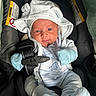 baby, infant, car_seat, blue_mittens, hooded_jacket, safety_belt, warning_label, cute, small, face, clothing, newborn, portrait, child, seat_belt, baby_clothes, indoor, closeup, expression, light