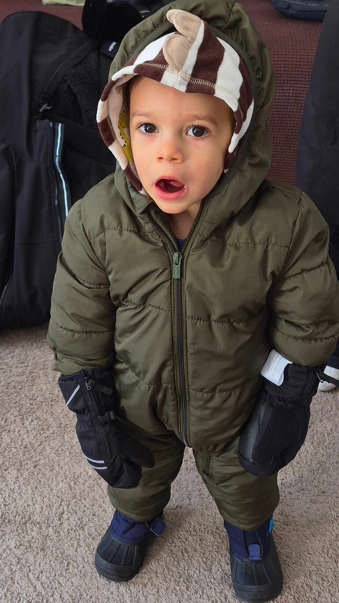 James is registered to the contest to win money with this photo: child, toddler, winter_clothing, snowsuit, mittens, hood, carpet, indoor, boots, curious, standing, brown_eyes, striped_hood, puffy_jacket, winter_gear, young_child, cute, warm_clothing, expression, casual