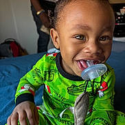 Nobel is registered to the contest to win money with this photo: toddler, child, pajamas, green_clothing, pacifier, toy, stuffed_animal, bed, blue_bedspread, smiling, happy, indoors, person, face, hair, hand, playful, blurred_background, adult, home