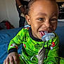 toddler, child, pajamas, green_clothing, pacifier, toy, stuffed_animal, bed, blue_bedspread, smiling, happy, indoors, person, face, hair, hand, playful, blurred_background, adult, home