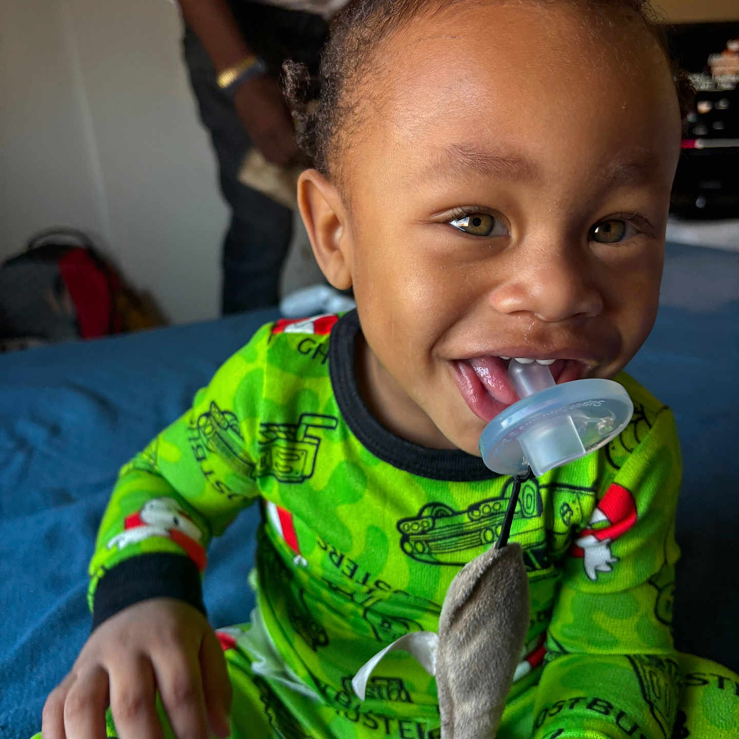 Nobel is registered to the contest to win money with this photo: adult, bed, blue_bedspread, blurred_background, child, face, green_clothing, hair, hand, happy, home, indoors, pacifier, pajamas, person, playful, smiling, stuffed_animal, toddler, toy