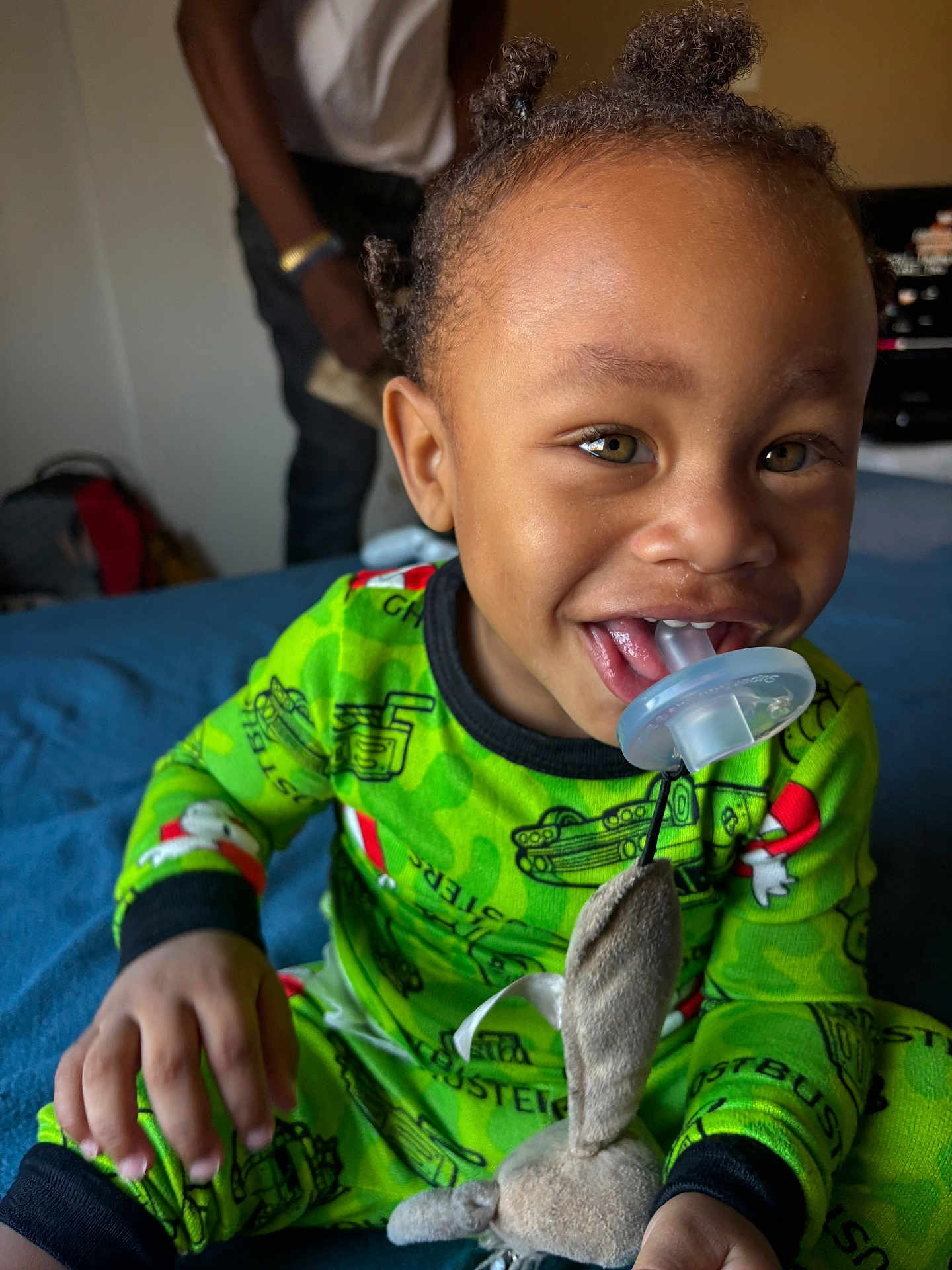 Nobel is registered to the contest to win money with this photo: toddler, child, pajamas, green_clothing, pacifier, toy, stuffed_animal, bed, blue_bedspread, smiling, happy, indoors, person, face, hair, hand, playful, blurred_background, adult, home
