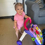 Allison is registered to the contest to win money with this photo: barefoot, child, colorful_toy, couch, face, floor, furniture, happy, home, indoor, person, pigtails, pillows, pink_clothing, play, smiling, standing, toddler, toy_walker, young_child