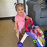 toddler, child, toy_walker, pigtails, pink_clothing, barefoot, floor, couch, pillows, indoor, smiling, face, person, play, home, furniture, colorful_toy, happy, young_child, standing