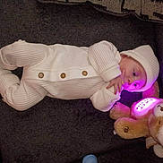 Samuel is registered to the contest to win money with this photo: baby, infant, newborn, knit_outfit, white_clothing, beanie, teddy_bear, plush_toy, glowing_light, couch, blanket, buttons, hand, face, eyes, lying_down, indoor, cozy, sleepwear, portrait