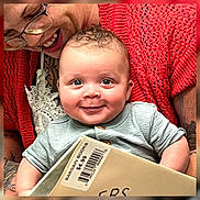 Bradley joined the competition — help win amazing prizes! baby, adult, book, smile, glasses, red_sweater, tattoo, curly_hair, holding, barcode, price_tag, indoor, closeup, portrait, cute, happy, person, face, child, family
