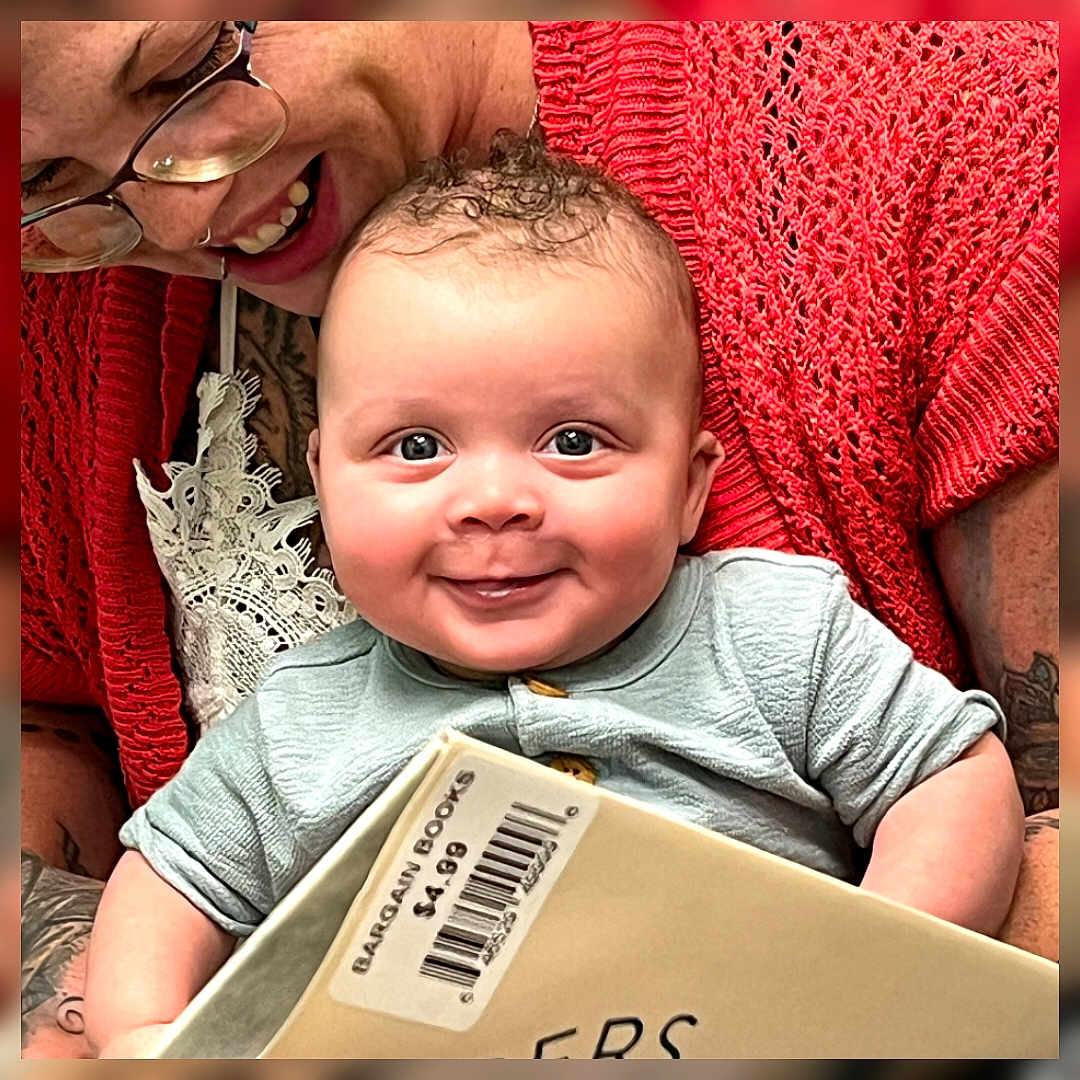 Bradley joined the competition — help win amazing prizes! baby, adult, book, smile, glasses, red_sweater, tattoo, curly_hair, holding, barcode, price_tag, indoor, closeup, portrait, cute, happy, person, face, child, family