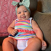 Charli is registered to the contest to win money with this photo: baby, child, infant, smiling, headband, bow, striped_dress, couch, indoors, cute, chubby_arms, chubby_legs, happy, portrait, relaxing, furniture, skin, person, seated, home
