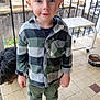 child, boy, smiling, standing, checkered_shirt, green_pants, spiderman_shoes, dog, dog_food_bowl, porch, tiles, railing, stone_wall, outdoor, casual_clothing, cute, young_child, pet, daylight, happy