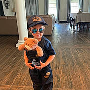 Anthony is registered to the contest to win money with this photo: child, teddy_bear, sunglasses, cap, denim_overalls, boots, living_room, wood_floor, sofa, curtains, table, chair, window, indoor, casual, person, toy, fashion, hat, home