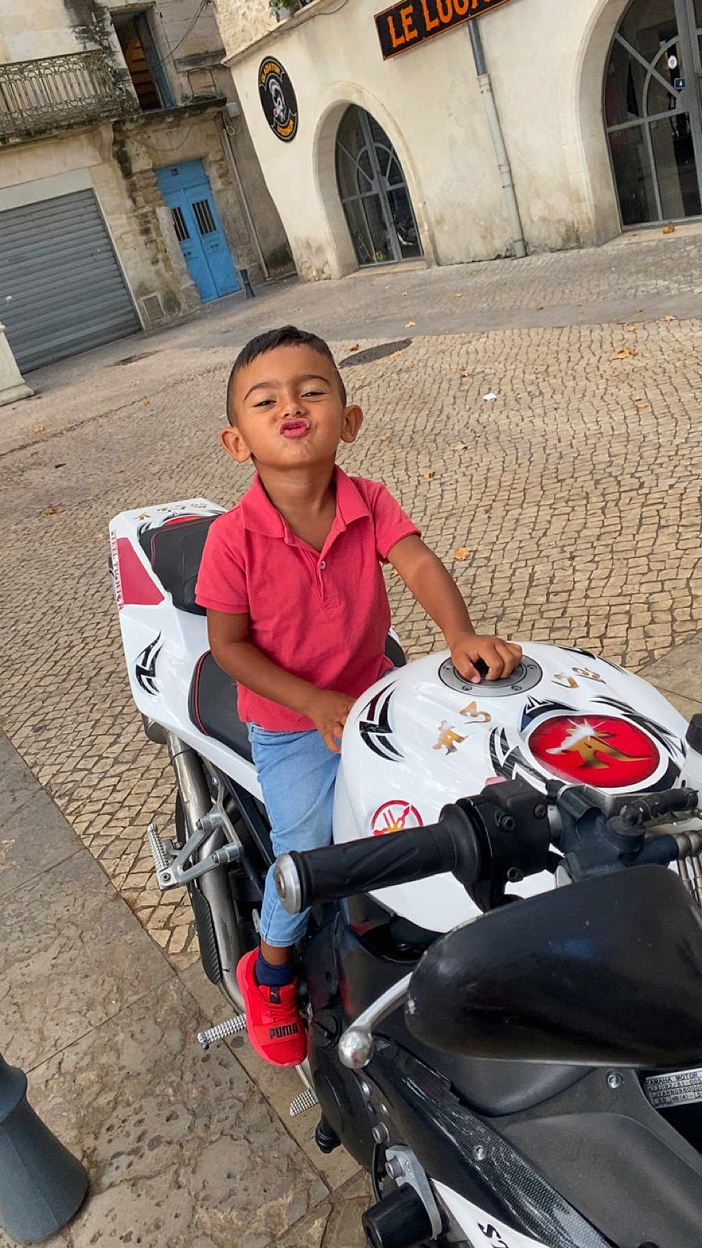 Ezai participe au concours pour gagner de l'argent avec cette photo : child, motorcycle, red_shoes, red_shirt, cobblestone, street, building, playful, kiss_face, outdoor, person, vehicle, sidewalk, puma, casual_clothing, young_child, urban, daylight, fun, transport