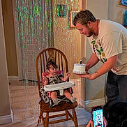 Hope joined the competition — help win amazing prizes! toddler, child, birthday_cake, high_chair, wooden_chair, man, woman, smartphone, streamers, curly_hair, indoor, celebration, candle, floor_tile, wall, dress, socks, photography, family, party