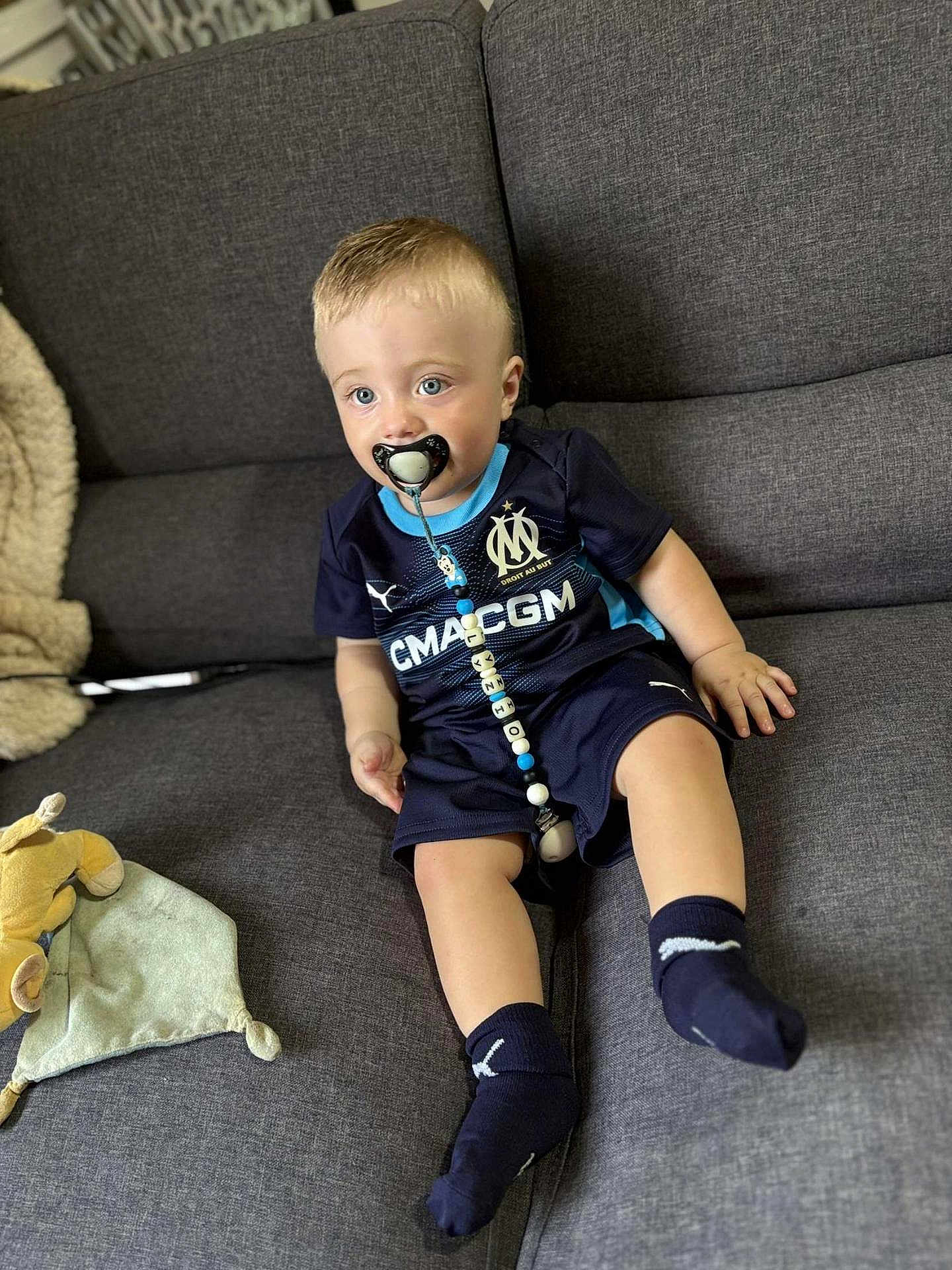 Lyzio participe au concours pour gagner de l'argent avec cette photo : toddler, pacifier, child, blue_eyes, sports_jersey, socks, couch, toy, blanket, indoor, person, seated, beads, clothing, furniture, cute, comfort, home, portrait, young_child