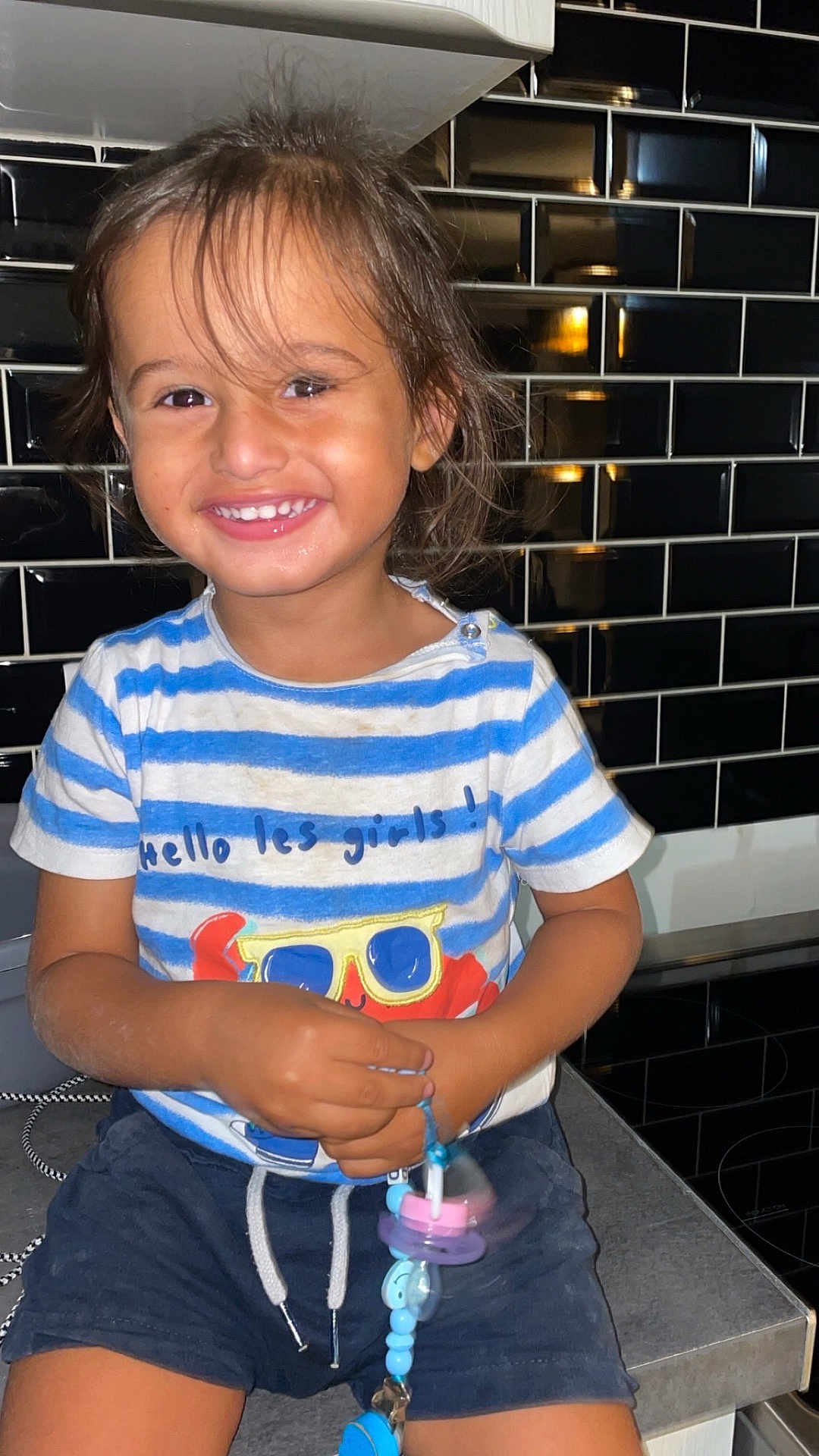 Farid participe au concours pour gagner de l'argent avec cette photo : toddler, child, smiling, happy, striped_shirt, crab_illustration, pacifier, kitchen, counter, black_tiles, shorts, indoor, cute, portrait, young_child, casual_clothing, playful, holding, seated, bright_lighting