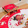 newborn, baby, sleeping, colorful_clothing, headband, traditional_attire, mat, floor, decorative_stool, hand, rings, poster, text, wall, wooden_floor, flower_pattern, animal_pattern, indoors, child, peaceful