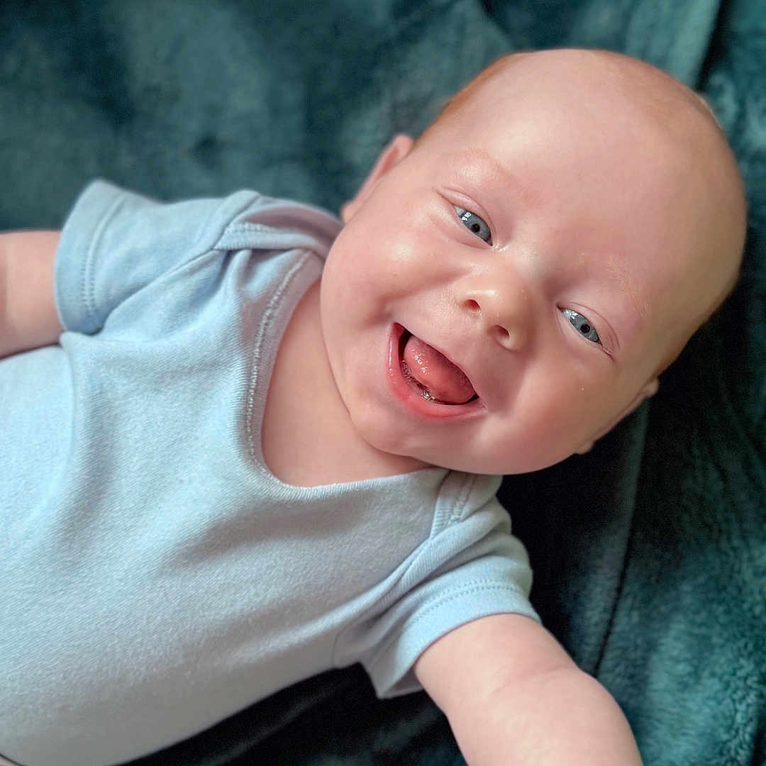 Toby is registered to the contest to win money with this photo: arm, baby, baby_boy, blanket, blue_onesie, child, closeup, comfort, cute, expression, face, happy, head, infant, lying_down, portrait, skin, smiling, soft_texture, tongue_out
