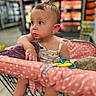 baby, shopping_cart, polka_dot, plush_toy, child, supermarket, grocery_store, curious, pink, clothing, seat_cover, infant, cute, portrait, indoor, shopping, person, toy, expression, blurred_background