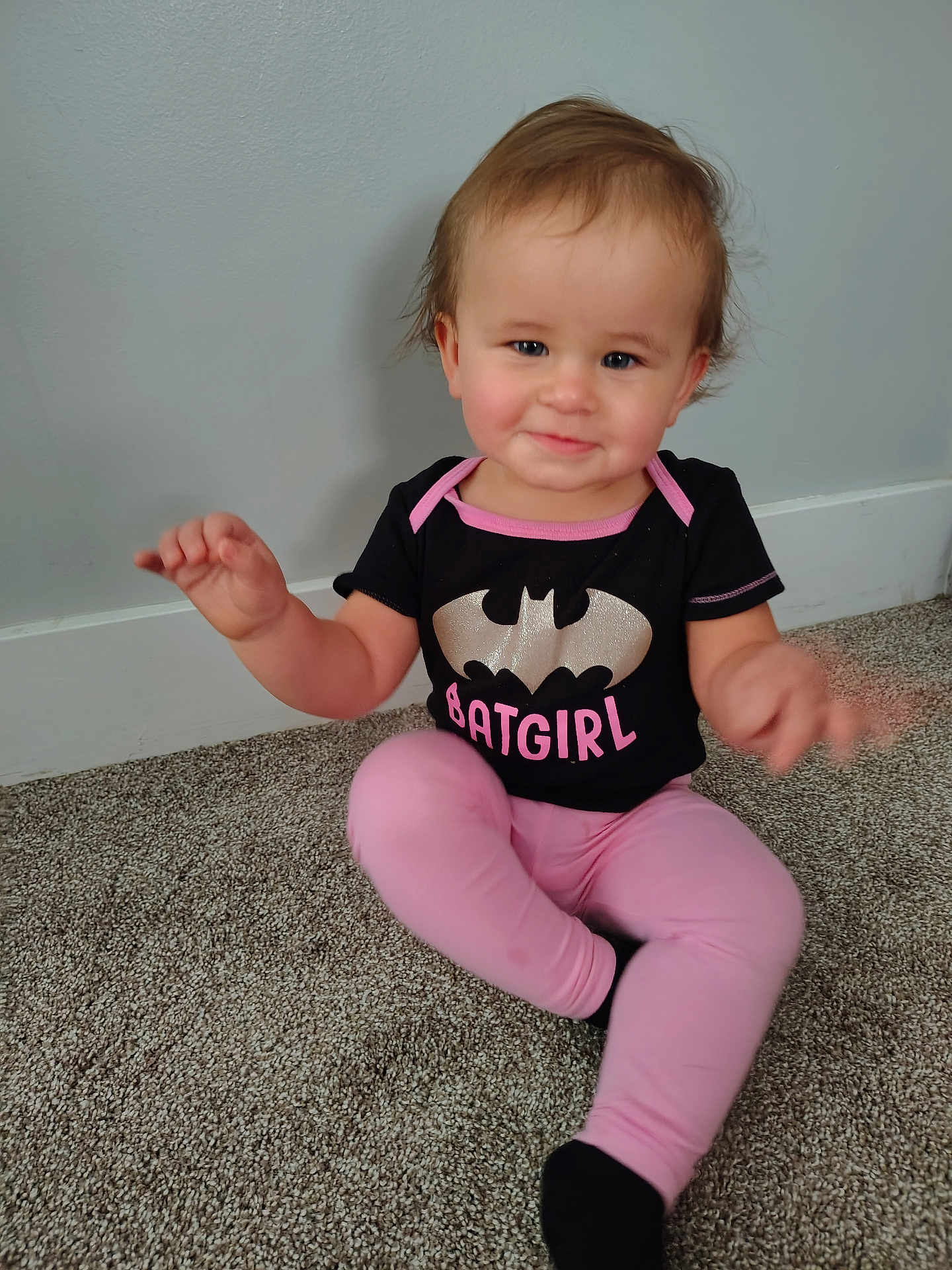 Bernadette joined the competition — help win amazing prizes! baby, child, infant, sitting, carpet, black_shirt, batgirl, pink_leggings, socks, smiling, indoor, person, cute, hair, toddler, wall, floor, clothing, playful, portrait