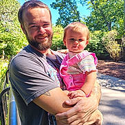 Bernadette is registered to the contest to win money with this photo: man, child, toddler, beard, smile, outdoor, sunlight, trees, greenery, clothing, pink_overalls, holding, happy, nature, daylight, casual, portrait, person, family, park
