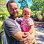 man, child, toddler, beard, smile, outdoor, sunlight, trees, greenery, clothing, pink_overalls, holding, happy, nature, daylight, casual, portrait, person, family, park