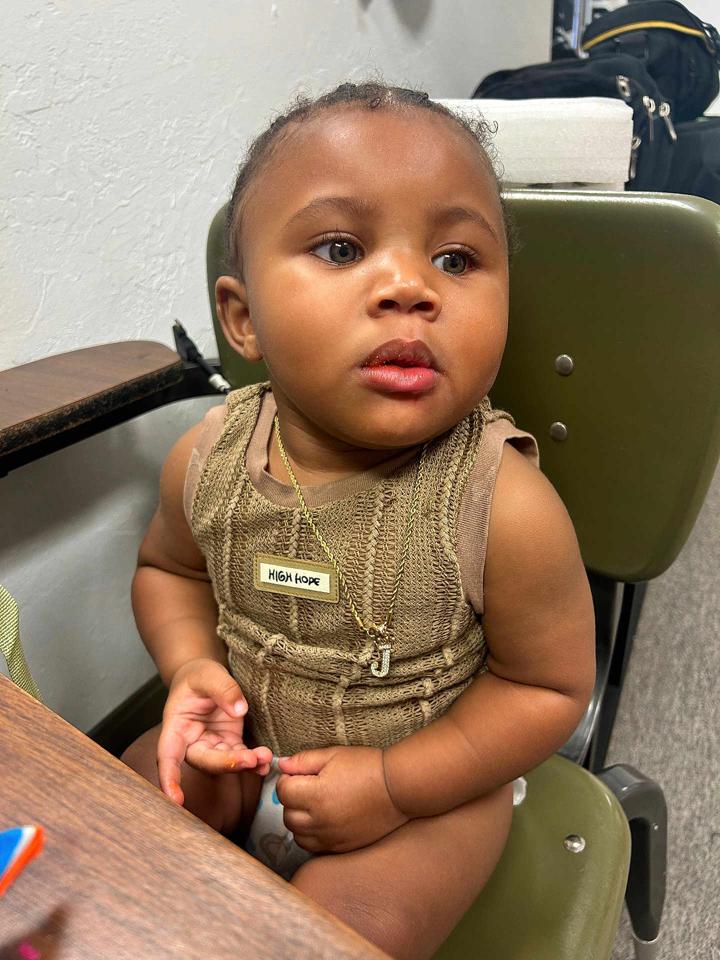 Jhamir is registered to the contest to win money with this photo: toddler, child, face, necklace, jewelry, chair, green_chair, brown_clothing, badge, indoor, table, curly_hair, skin, fingers, expression, young_child, person, seated, looking_away, cute