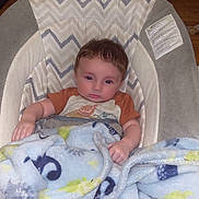 Xavier is registered to the contest to win money with this photo: baby, baby_chair, blanket, child, clothing, comfort, curly_hair, face, floor, foot, hand, home, indoor, infant, looking_at_camera, pattern, relaxed, seated, soft_texture, wood_floor