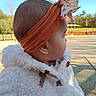 baby, bow, cheek, child, close_up, coat_button, ear, earring, floral_bow, fluffy_coat, headband, infant, orange_headband, outdoor, park, portrait, profile, sky, trees, white_jacket