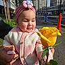 adult_hand, baby, buildings, child, city_street, flower, girl, hand, headband, infant, knitted_cardigan, outdoors, pink_headband, portrait, sidewalk, smiling, sunlight, sweater, urban, yellow_rose