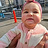 adult_hands, baby, building, cardigan, child, chubby_cheeks, close_up, earrings, hands, headband, infant, knit_sweater, outdoor, pink_headband, portrait, smiling, street, sunlight, traffic_cone, urban