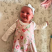 Bonnie is registered to the contest to win money with this photo: baby, infant, smiling, headband, pink_romper, strawberry_pattern, white_long_sleeve, lying_down, blanket, plush_toy, cute, happy, child, indoor, soft_toys, patterned_background, portrait, closeup, cozy, baby_clothing