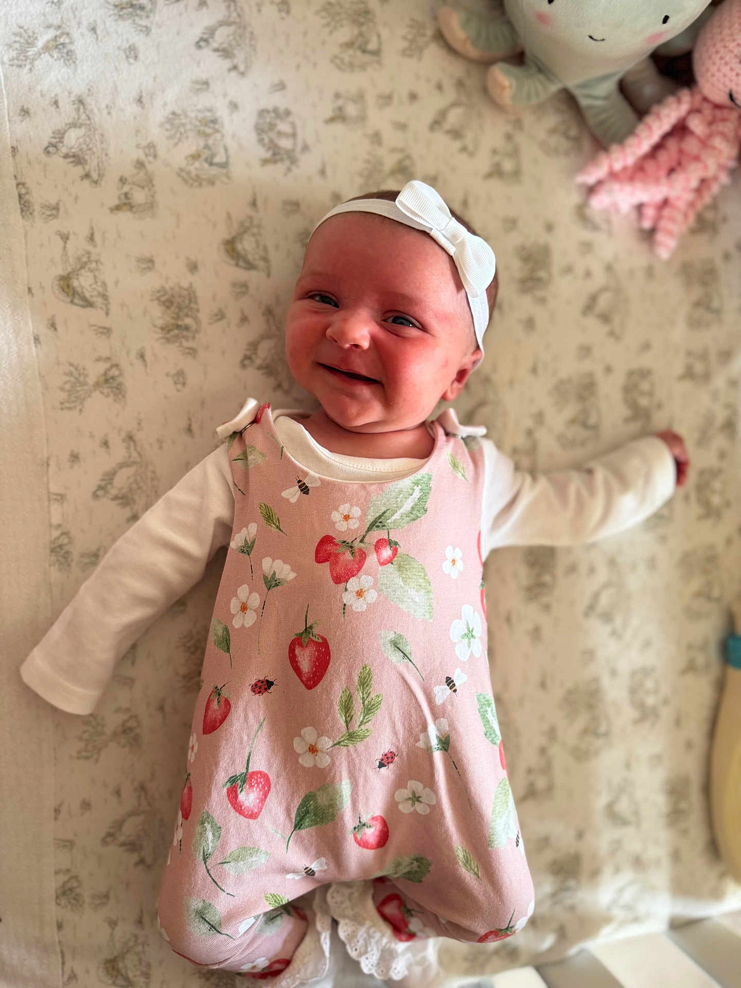 Bonnie is registered to the contest to win money with this photo: baby, infant, smiling, headband, pink_romper, strawberry_pattern, white_long_sleeve, lying_down, blanket, plush_toy, cute, happy, child, indoor, soft_toys, patterned_background, portrait, closeup, cozy, baby_clothing