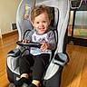toddler, child, car_seat, indoor, wooden_floor, smiling, steering_wheel, black_pants, gray_shirt, socks, hair, face, furniture, playpen, home, happy, seated, toy, person, room