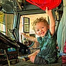 child, curly_hair, smiling, vehicle_interior, driver_seat, red_object, steering_wheel, window, sunlight, dashboard, controls, seat, casual_clothing, happy, playful, indoor, young_child, excited, transportation, cabin