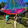 woman, dog, grass, outdoor, people, colorful, ribbons, canopy, sunny, blue_sky, trees, casual_clothing, smiling, holding, nature, festival, shadows, park, event, daylight