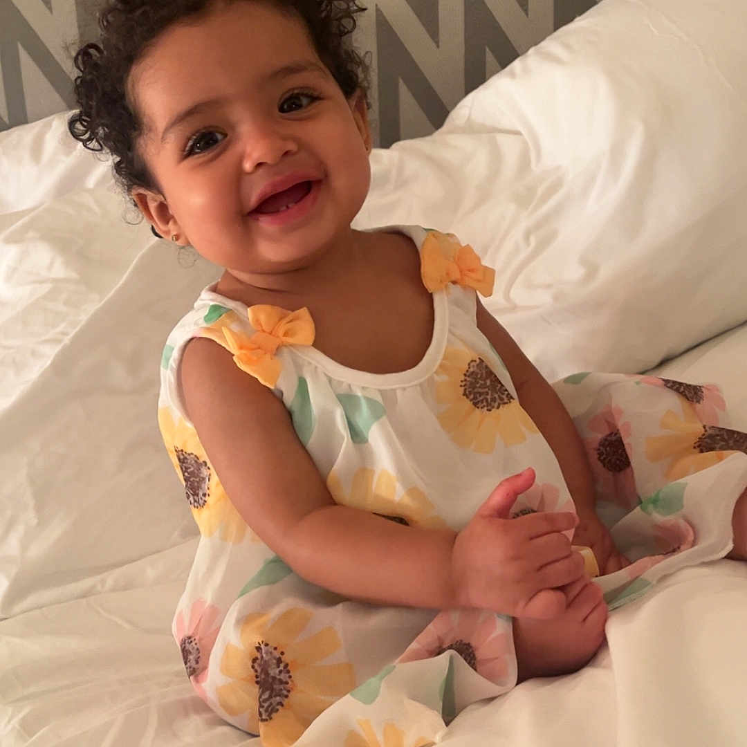 Layla is registered to the contest to win money with this photo: baby, bed, child, curly_hair, cute, dress, fashion, floral_pattern, happy, home, indoor, person, portrait, sitting, skin, smiling, toddler, white_bedding, yellow_bows, zigzag_wall