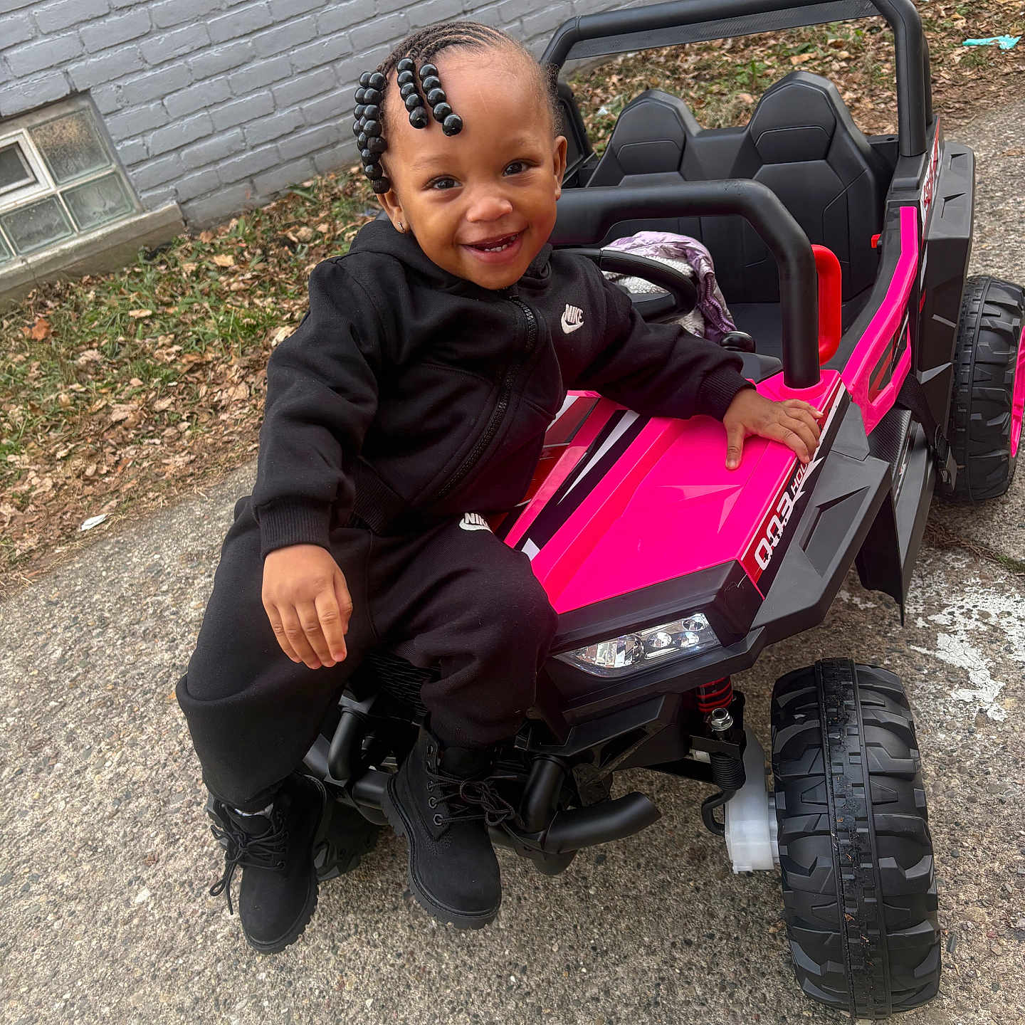 Kennedi is registered to the contest to win money with this photo: toddler, child, smiling, toy_car, pink, black_clothing, nike, outdoor, pavement, brick_wall, beaded_hair, boots, happy, person, play, vehicle, small_car, leaves, casual, daylight