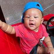 Ryaire joined the competition — help win amazing prizes! baby, child, red_shirt, blue_cap, toy_car, smiling, indoor, seatbelt, playful, happy, person, vehicle, child_seat, floor, earrings, hand, face, headwear, casual_clothing, infant