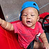 baby, child, red_shirt, blue_cap, toy_car, smiling, indoor, seatbelt, playful, happy, person, vehicle, child_seat, floor, earrings, hand, face, headwear, casual_clothing, infant