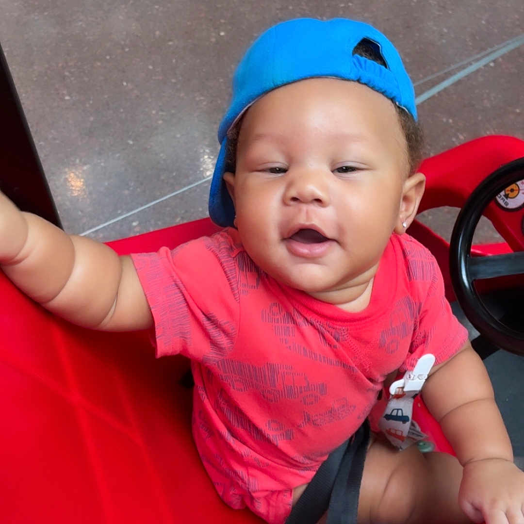 Ryaire joined the competition — help win amazing prizes! baby, child, red_shirt, blue_cap, toy_car, smiling, indoor, seatbelt, playful, happy, person, vehicle, child_seat, floor, earrings, hand, face, headwear, casual_clothing, infant