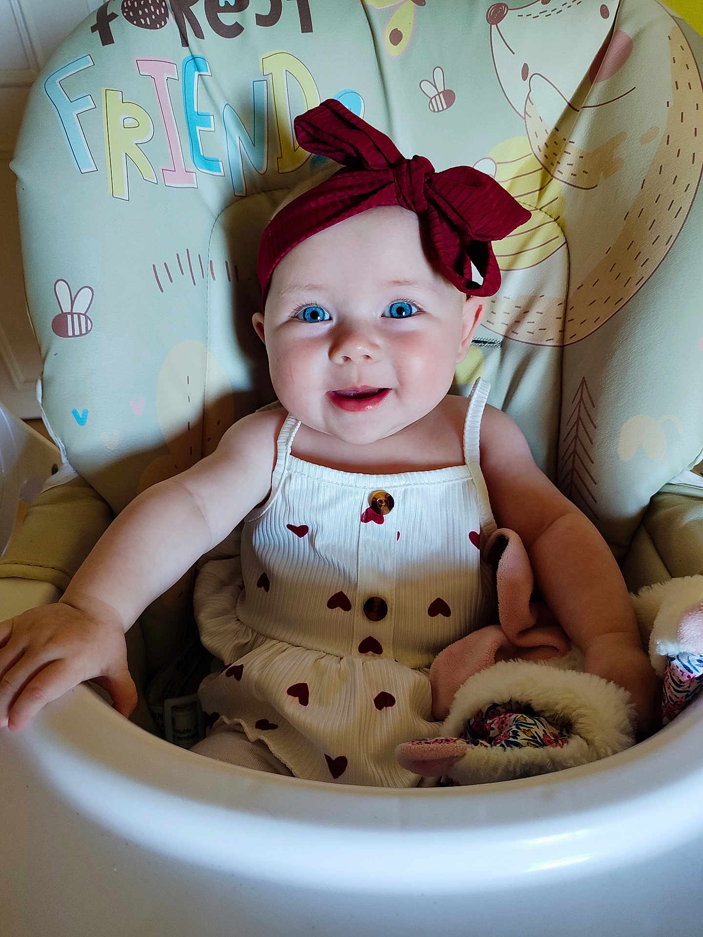 Lilwenn participe au concours pour gagner de l'argent avec cette photo : baby, child, high_chair, headband, blue_eyes, smiling, dress, heart_pattern, toy, soft_toy, indoor, person, infant, cute, happy, seated, baby_clothing, face, portrait, decorative_background