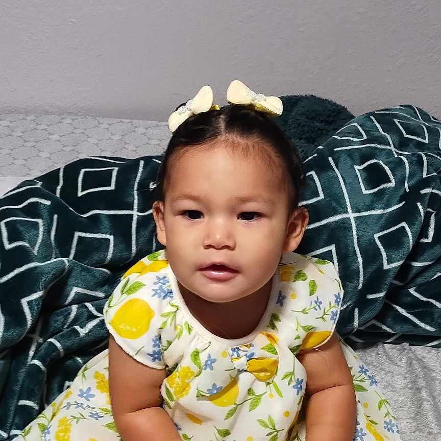 Rebecca is registered to the contest to win money with this photo: toddler, child, girl, dress, hairbrush, hair_bow, bed, blanket, pattern, yellow, white, blue, green, foot, sitting, indoors, curious, person, portrait, cute