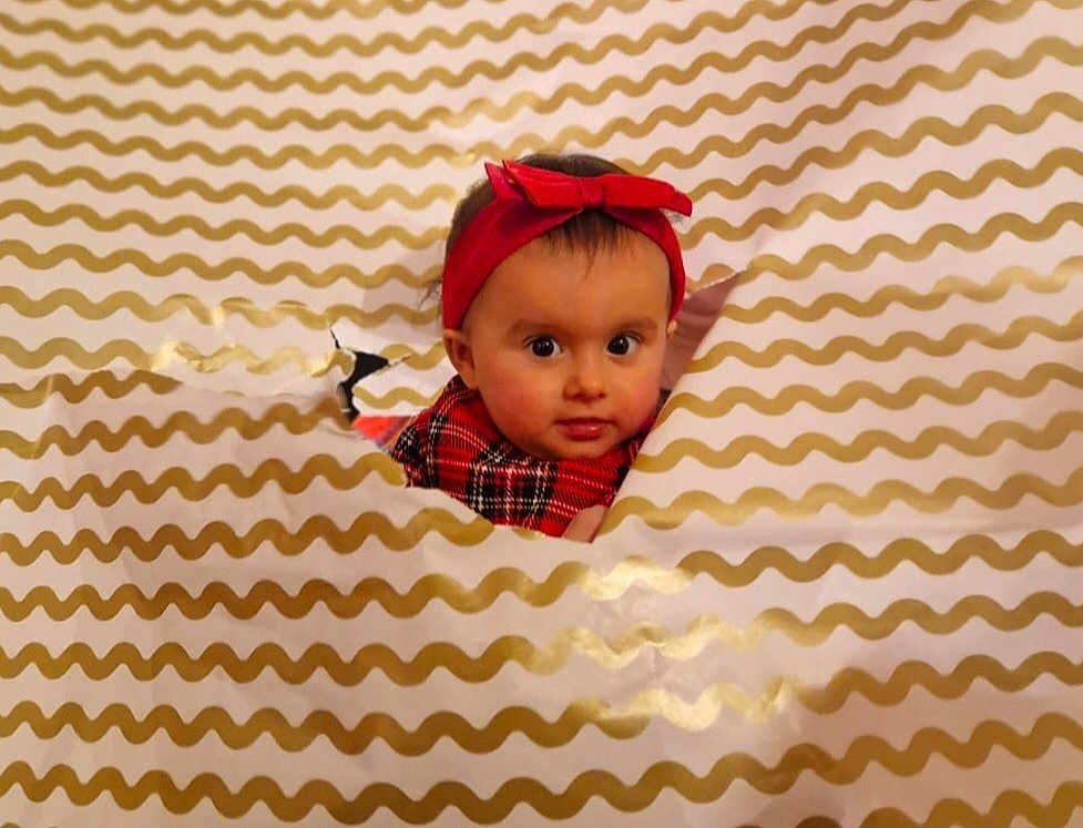Nina participe au concours pour gagner de l'argent avec cette photo : baby, child, face, headband, red, plaid, pattern, wrapping_paper, hole, curious, peek, indoor, cute, portrait, infant, expression, holiday, festive, smiling, young