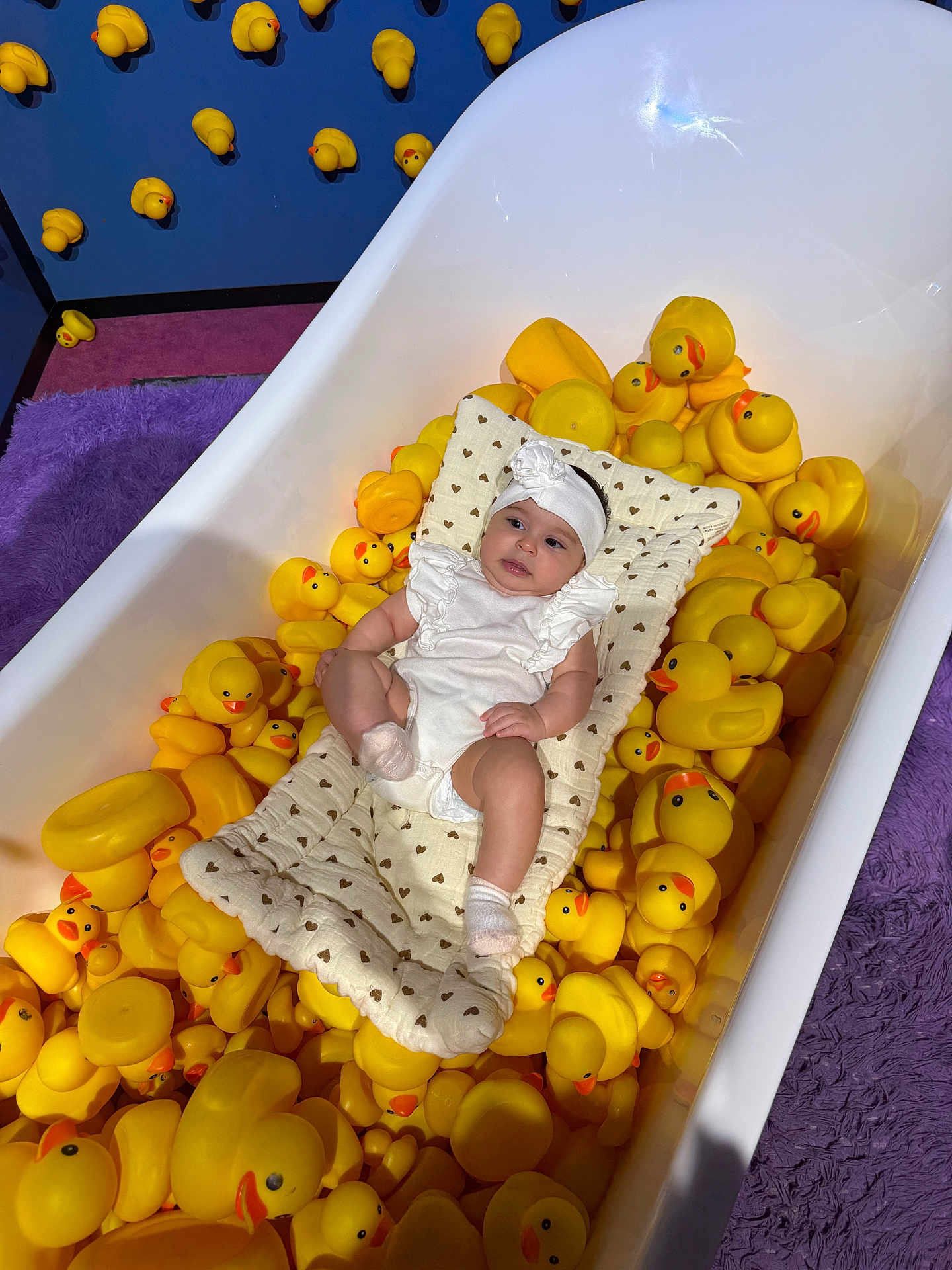 Mary joined the competition — help win amazing prizes! baby, bathtub, rubber_duck, yellow, cushion, white_clothing, headband, purple_rug, pink_rug, blue_wall, toy, child, indoor, playful, cute, infant, soft_texture, patterned_cushion, relaxed, colorful