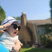 Uriel is registered to the contest to win money with this photo: baby, blurred_background, building, casual, child, daylight, grass, greenery, hand, happy, hat, holding, outdoor, person, portrait, skin, smiling, sunglasses, sunny, white_clothing